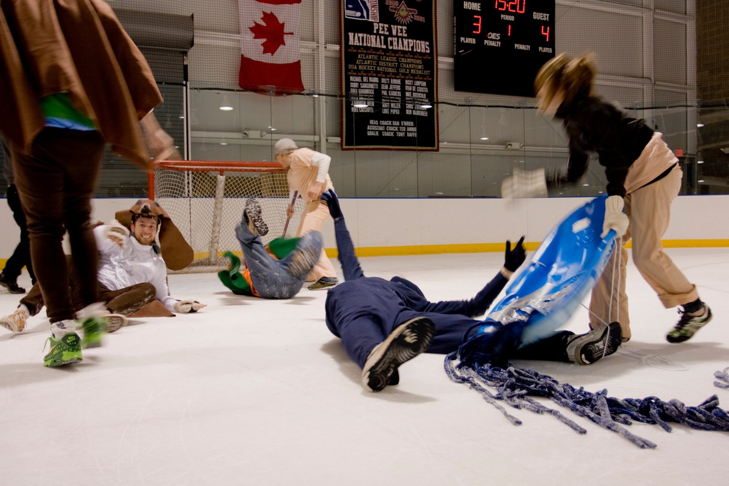 People in costumes sprawl and slide across an indoor ice rink, one pulling a blue sled, with a hockey goal and scoreboard behind them