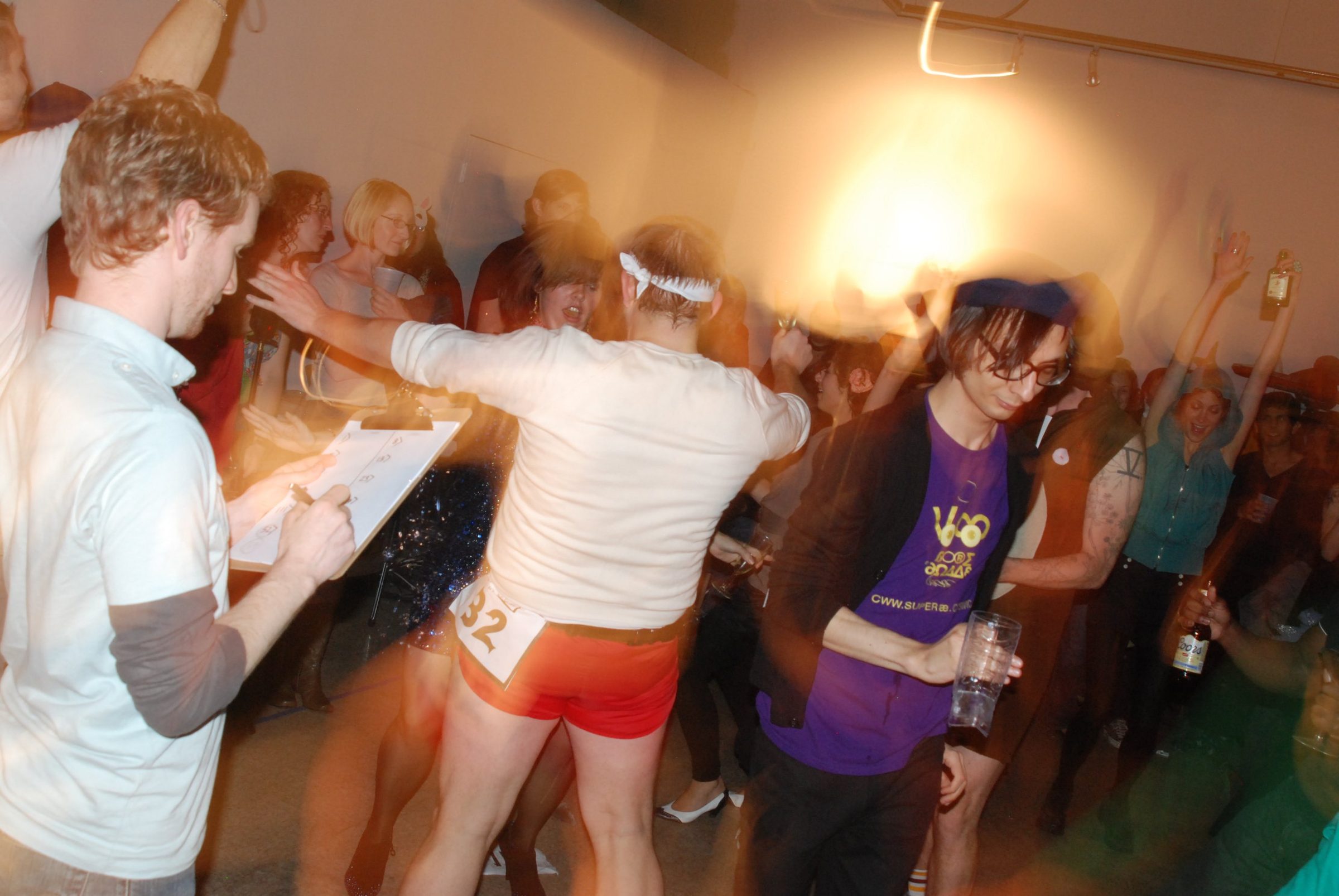 A crowd dancing with drinks in a gallery, one dancer in red shorts with a race number pinned on, while a man in a white shirt scores on a clipboard