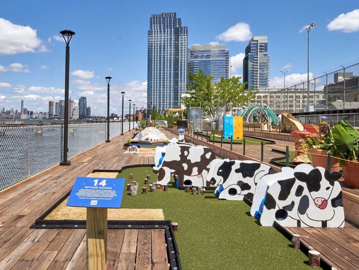 Cow-shaped mini golf obstacles on a green course along a wooden pier, with a numbered sign and the Manhattan skyline across the water