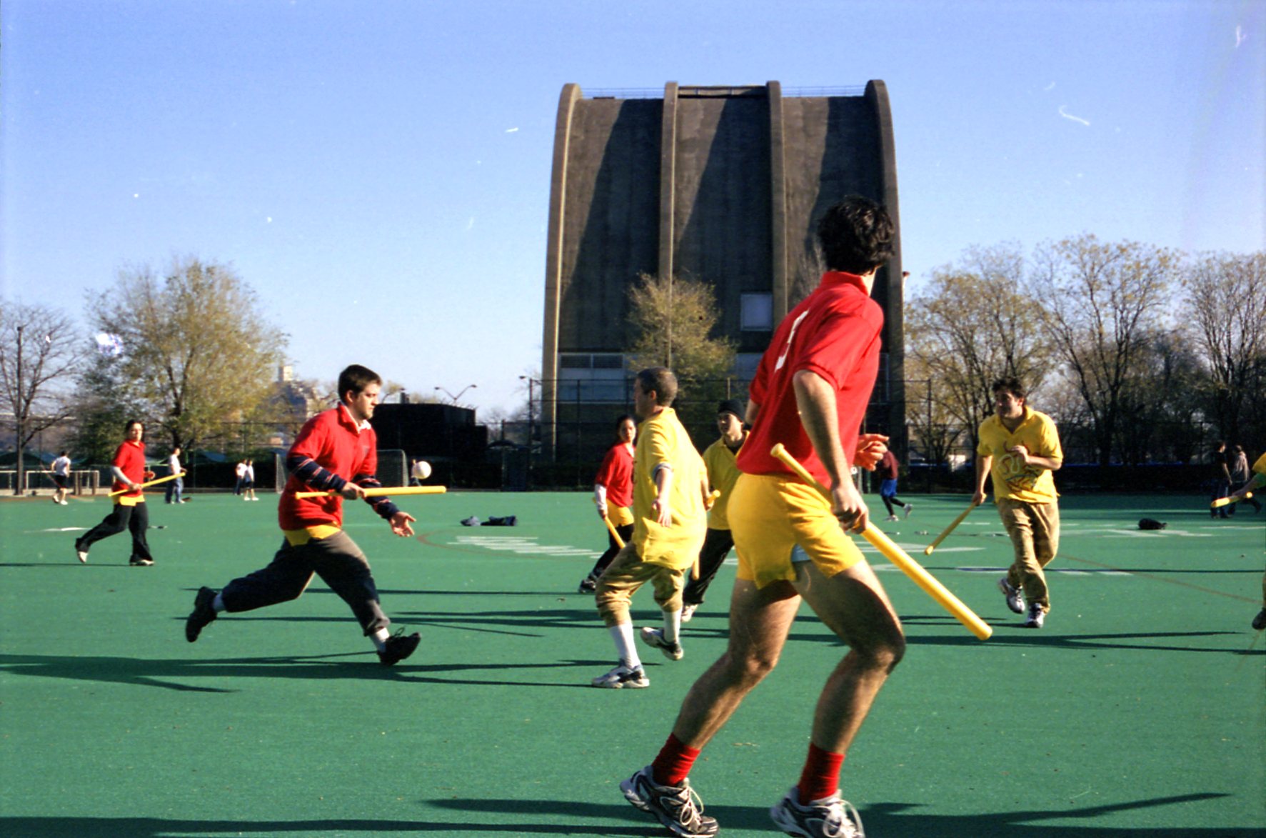 Players in red and yellow shirts run across a green field holding yellow wiffle bats, with a ball in the air, in front of a large concrete building and bare trees